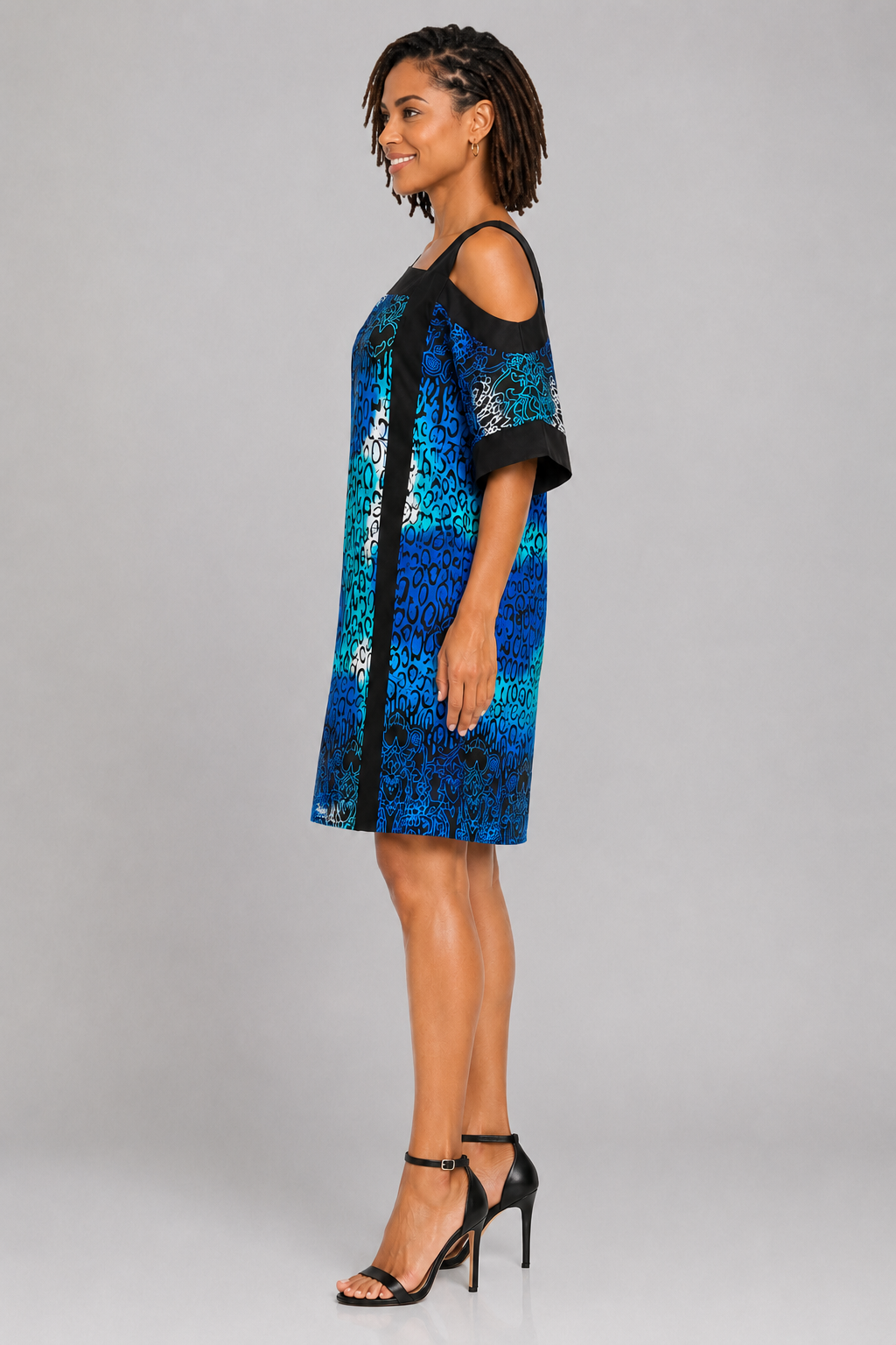 A woman modeling a short, cold-shoulder dress with a blue and black abstract pattern, paired with black high-heeled sandals, against a solid gray studio background.