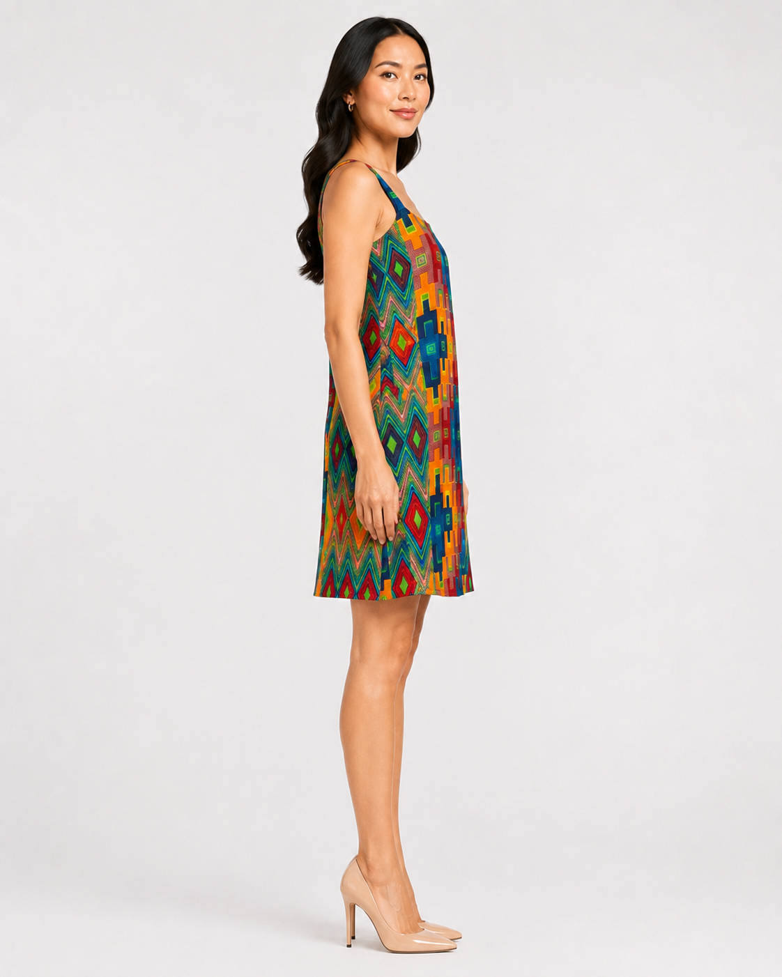 A woman modeling a sleeveless, V-neck shift dress with a vibrant multicolor geometric ikat pattern, paired with nude high heels, against a plain light gray studio background.