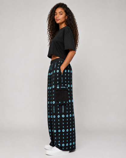 A woman stands in profile wearing a black cropped short-sleeve top, wide-leg black pants with a light blue geometric pattern and a side pocket, and white sneakers against a light gray studio background.