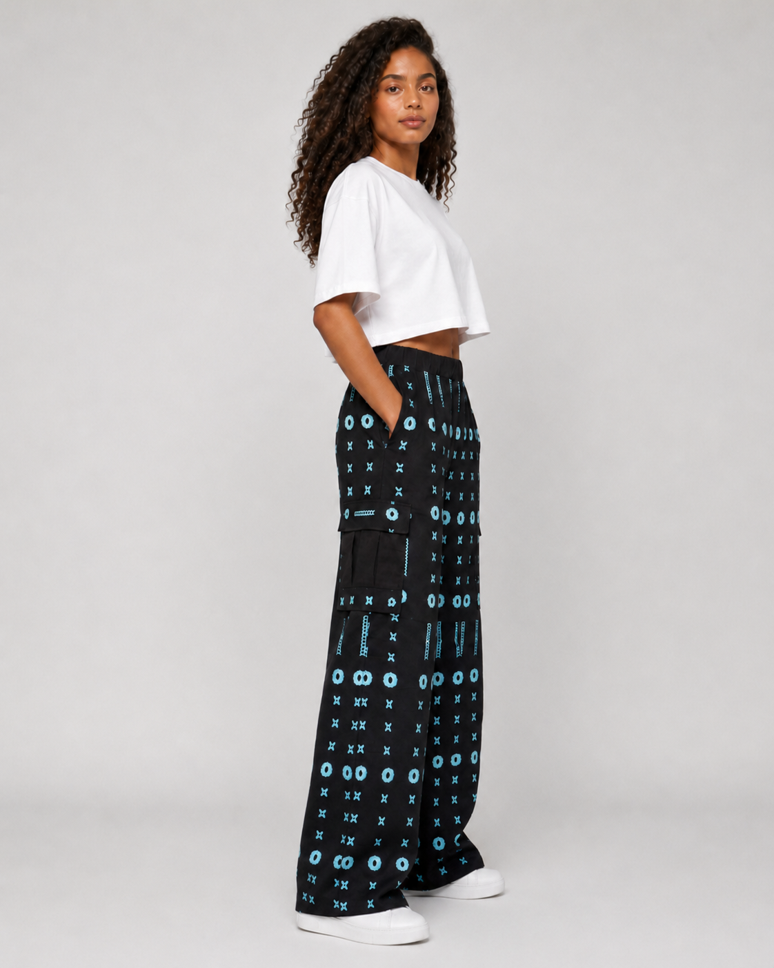 A model wearing black wide-leg pants with a blue and white geometric pattern, styled with a white cropped t-shirt and white sneakers, photographed against a light gray studio background.