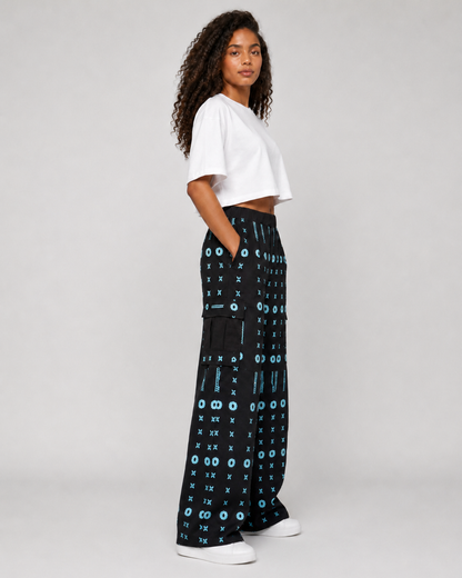A model wearing black wide-leg pants with a blue and white geometric pattern, styled with a white cropped t-shirt and white sneakers, photographed against a light gray studio background.