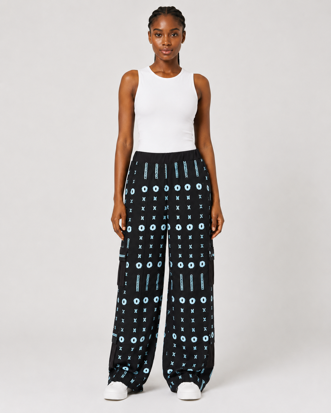 A model stands against a light gray studio background wearing black wide-leg pants with a light blue geometric pattern, a white sleeveless top, and white sneakers.
