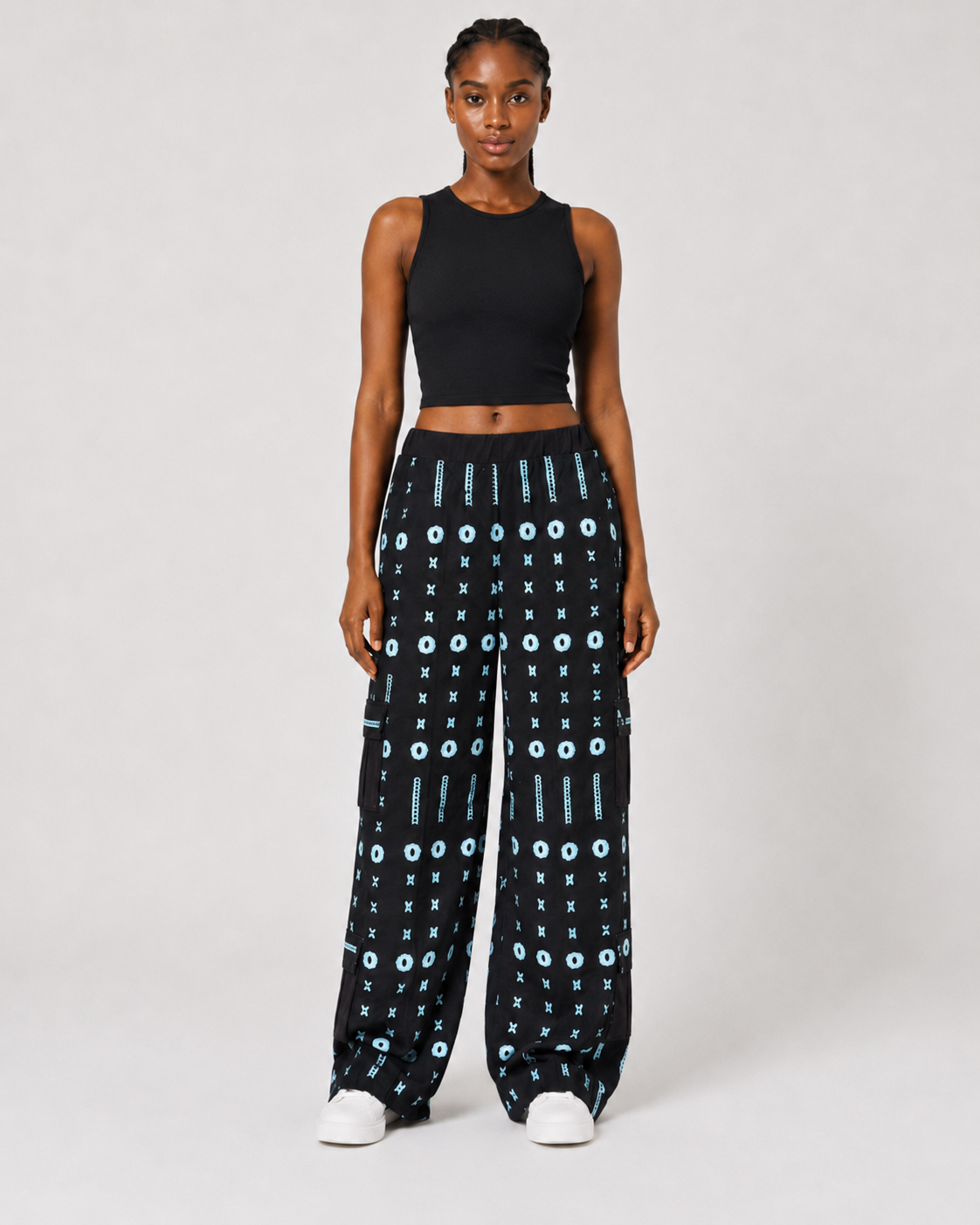 A model stands against a light gray studio background wearing a black sleeveless crop top and black wide-leg pants with a light blue geometric pattern, paired with white sneakers.
