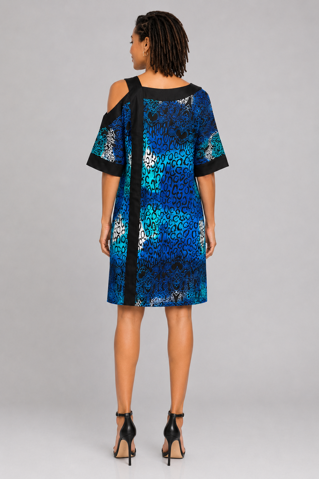 A woman viewed from the back wearing a short blue and black patterned dress with cold-shoulder sleeves and black trim, paired with black high-heeled sandals, against a light gray studio background.