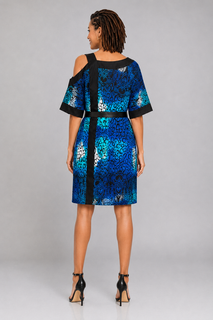 A woman viewed from the back wearing a short blue and black patterned dress with cold-shoulder sleeves and a matching black belt, standing against a light gray studio background.
