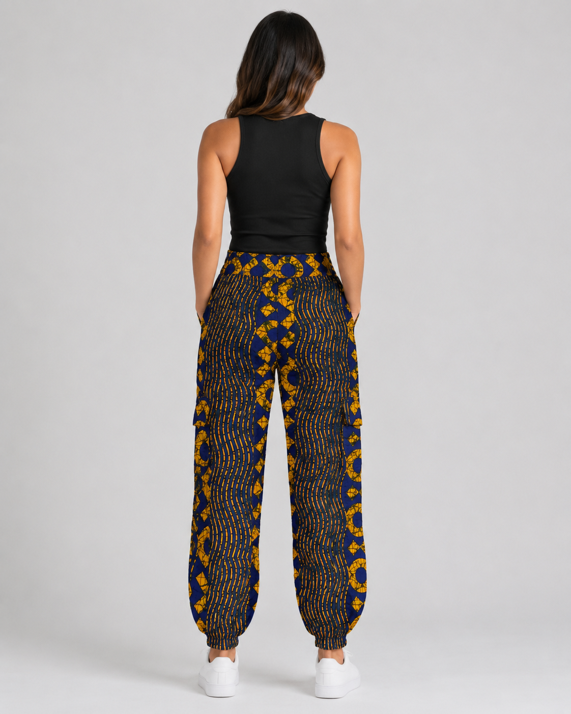 A woman shown from the back wearing loose, patterned pants with a black top and white sneakers, photographed against a plain light gray studio background.