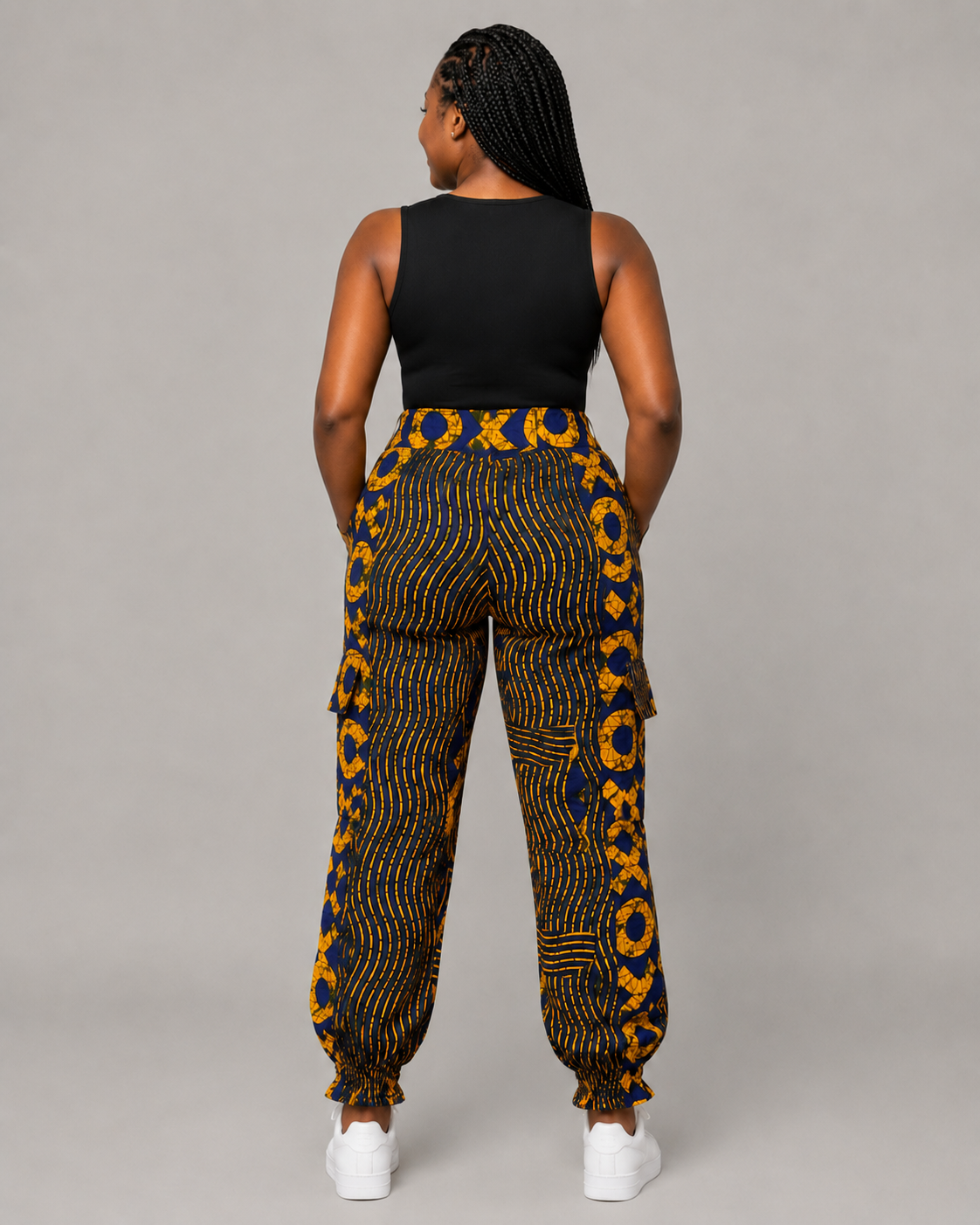 A woman shown from the back wearing high-waisted cargo pants with a bold yellow and blue pattern, a black sleeveless top, and white sneakers against a solid gray studio background.