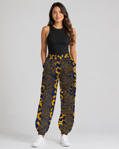 A woman modeling high-waisted, wide-leg pants with a bold yellow and navy blue wavy pattern, paired with a black sleeveless top and white sneakers, against a light gray studio background.