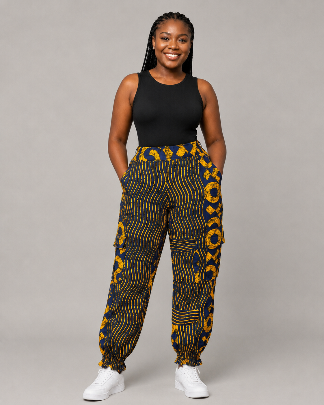 A woman modeling high-waisted, loose-fit pants with a bold yellow and navy blue African wax print, paired with a black tank top and white sneakers, against a solid gray studio background.