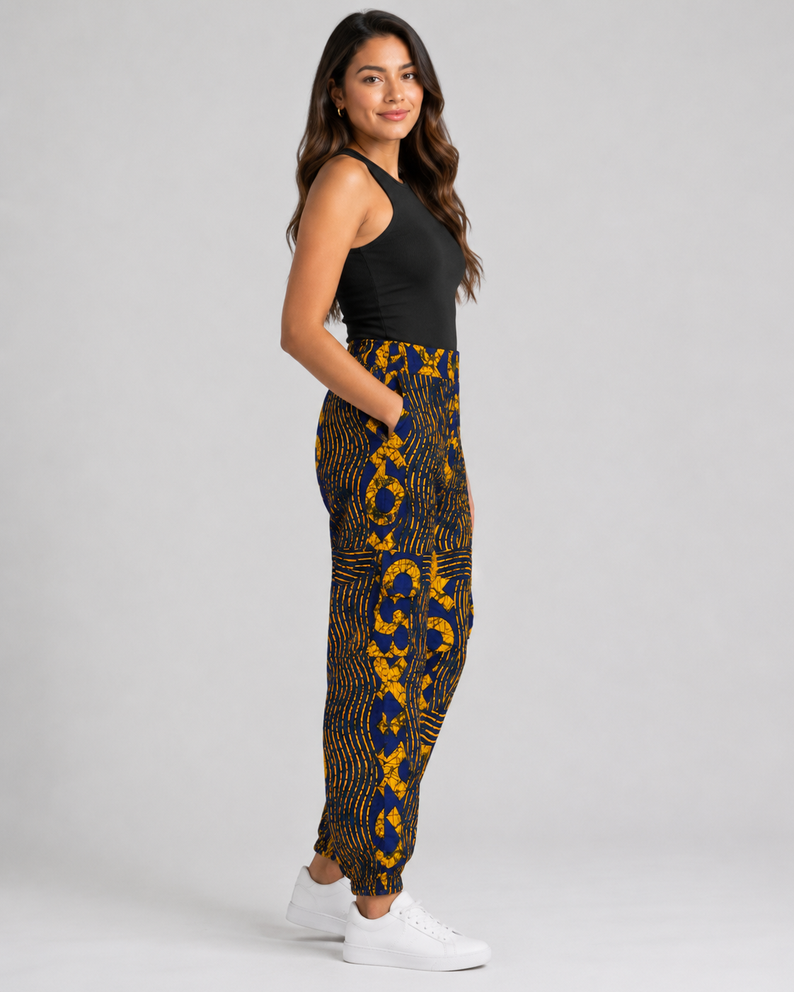 A woman modeling high-waisted, ankle-length pants with a bold yellow and navy blue abstract pattern, paired with a black sleeveless top and white sneakers, against a light gray studio background.