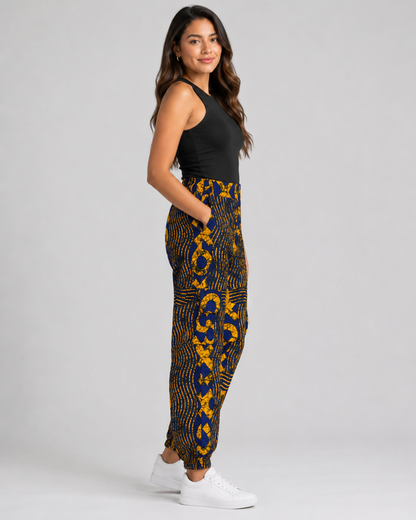 A woman modeling high-waisted, ankle-length pants with a bold yellow and navy blue abstract pattern, paired with a black sleeveless top and white sneakers, against a light gray studio background.