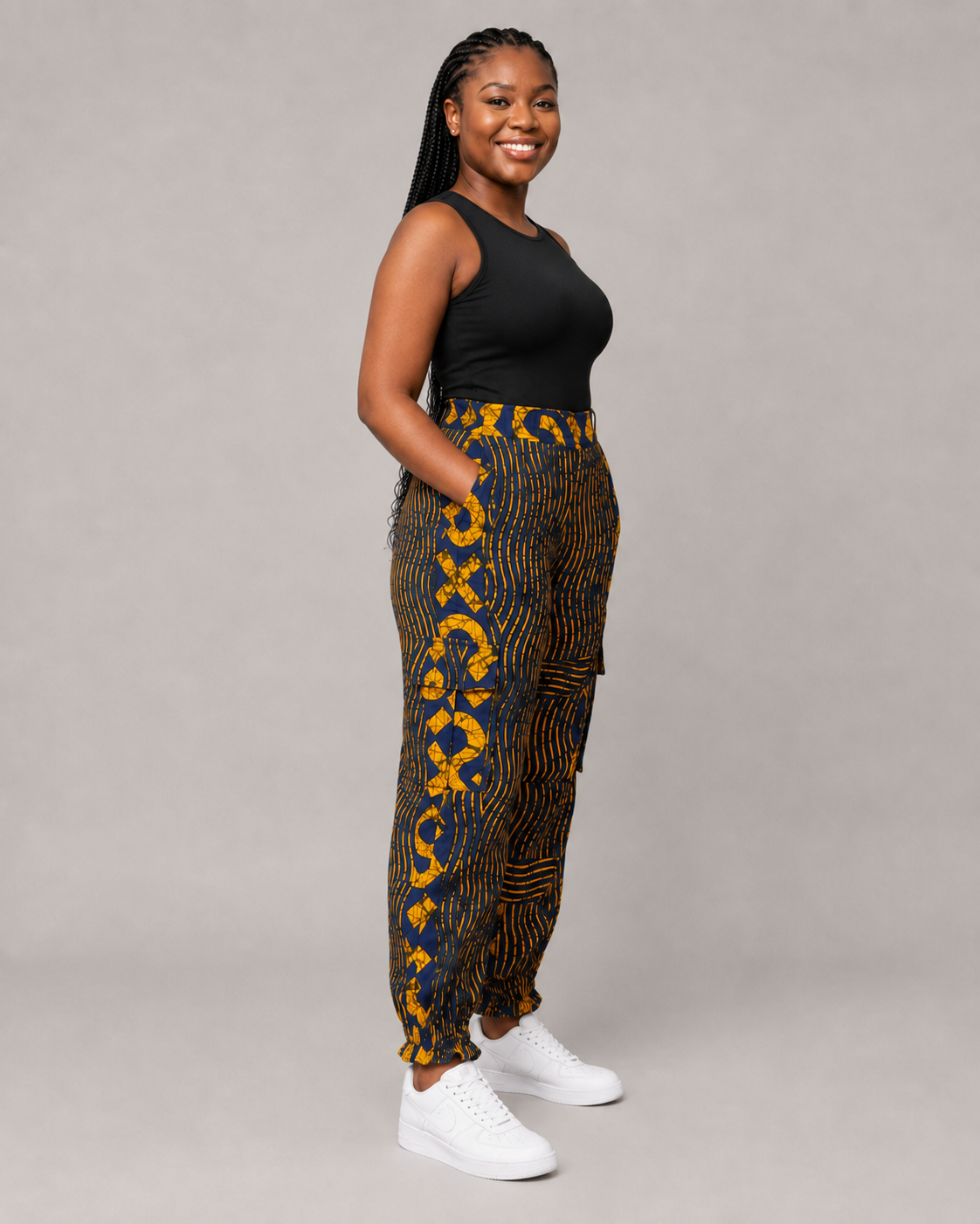 A woman modeling high-waisted, wide-leg pants with a bold yellow and dark blue pattern, paired with a black tank top and white sneakers, against a solid gray studio background.