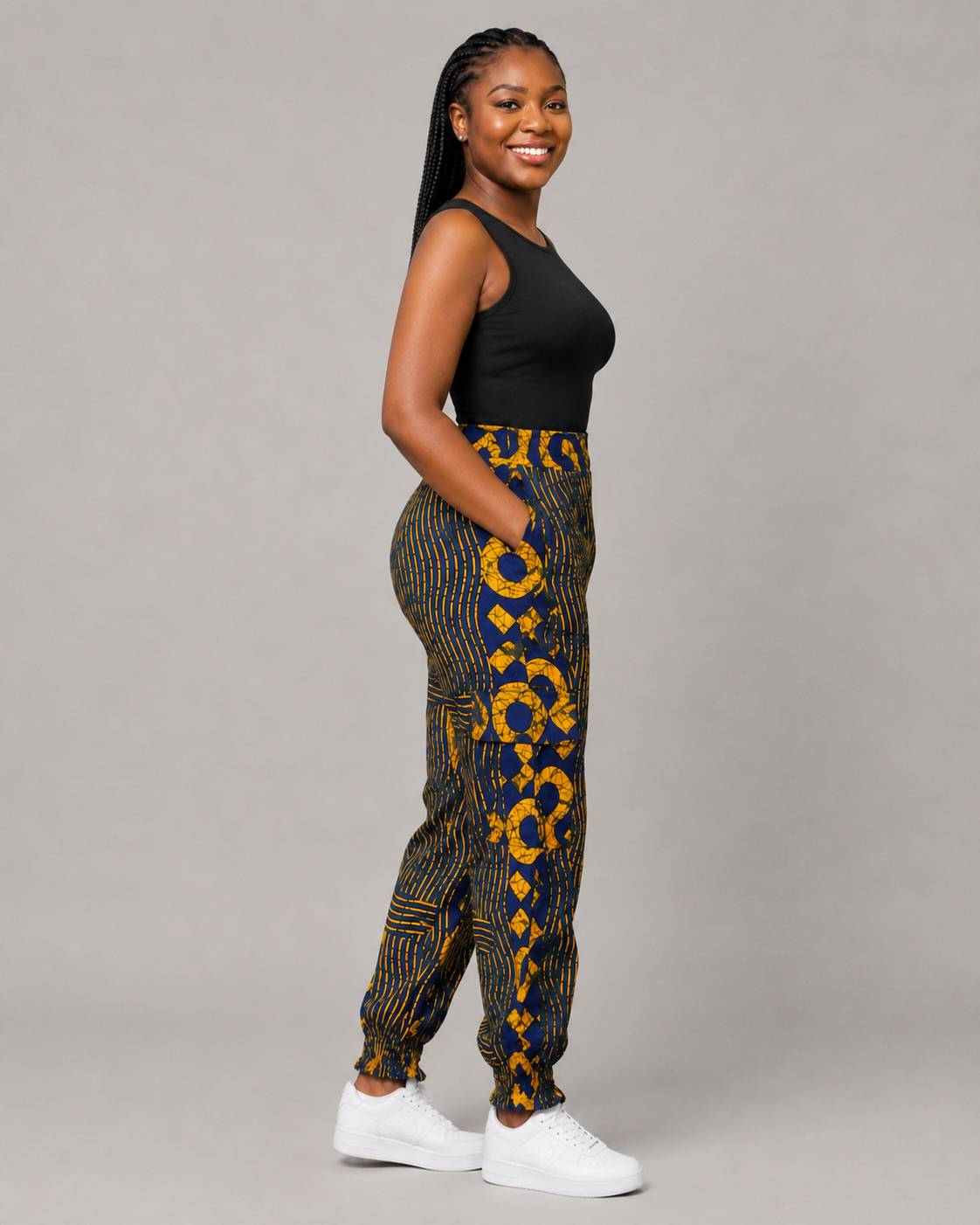 A woman modeling high-waisted, wide-leg pants with a bold yellow and black African wax print pattern, paired with a black sleeveless top and white sneakers, against a solid gray studio background.
