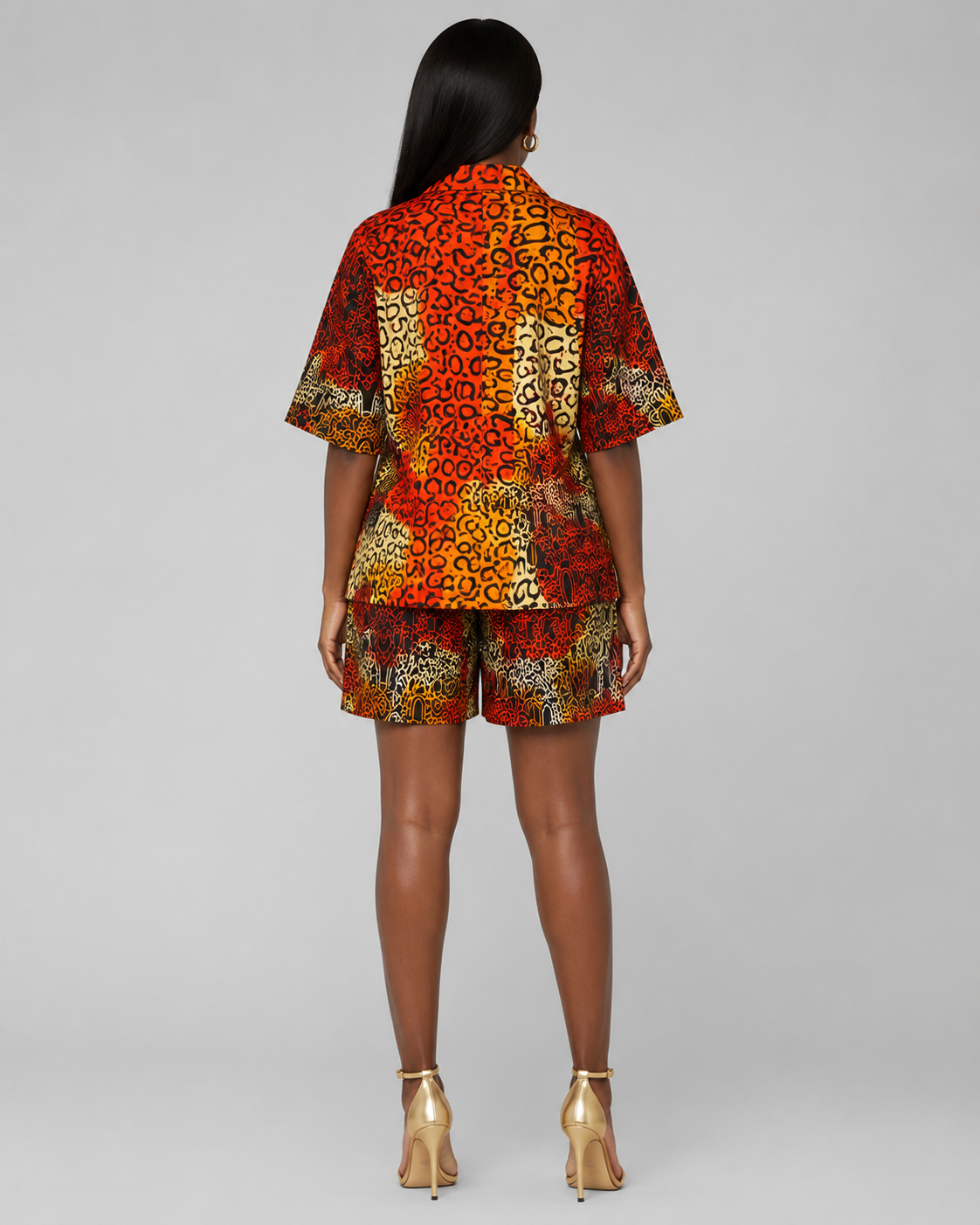 A model viewed from the back wearing a matching short-sleeve button-up shirt and shorts set with a vibrant red, orange, and yellow geometric pattern, paired with gold high-heeled sandals on a light gray background.