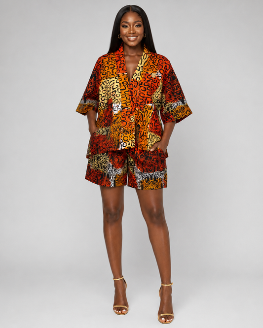 A woman modeling a matching two-piece outfit consisting of a short-sleeve button-front top and shorts, both featuring a bold multicolor abstract print in red, orange, yellow, black, and white, paired with gold high-heeled sandals against a light gray studio background.