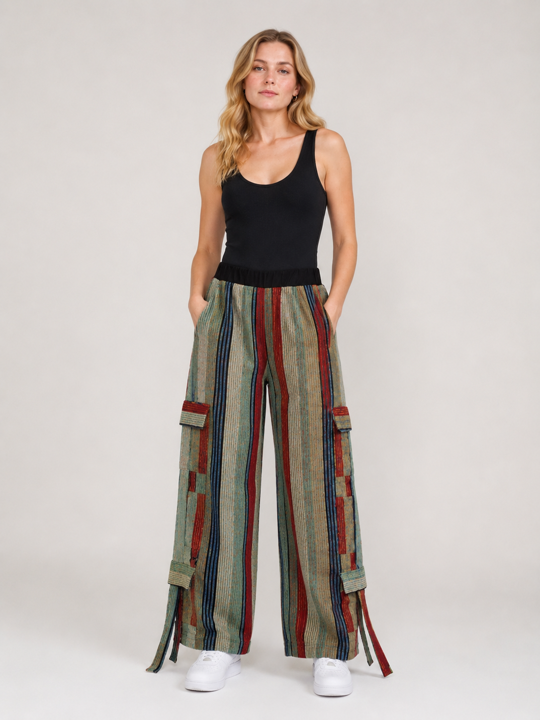 A woman modeling wide-leg cargo pants with vertical stripes in beige, green, red, and blue, paired with a black tank top and white sneakers, against a light gray studio background.
