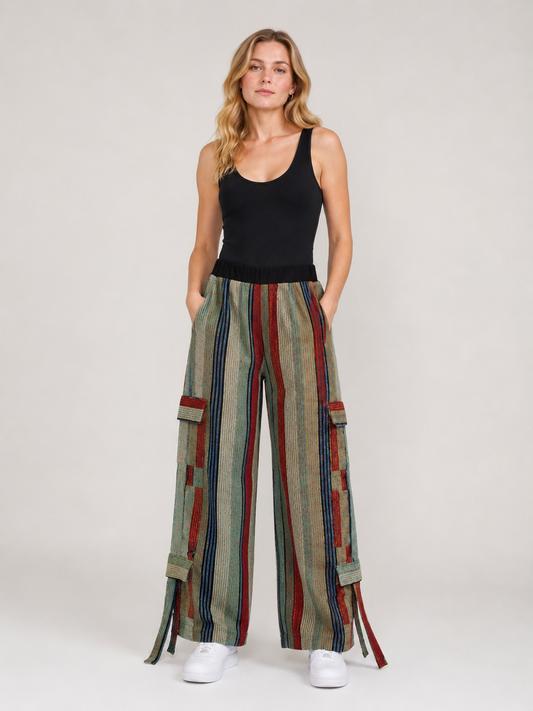 A woman modeling wide-leg cargo pants with vertical stripes in beige, green, red, and blue, paired with a black tank top and white sneakers, against a light gray studio background.