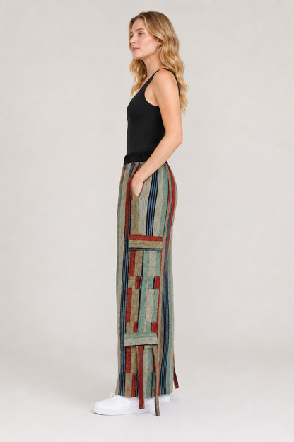 A woman stands in profile wearing a black sleeveless top and long, wide-leg trousers with a multicolor patchwork stripe pattern in muted red, blue, green, and beige tones, against a plain light gray studio background.