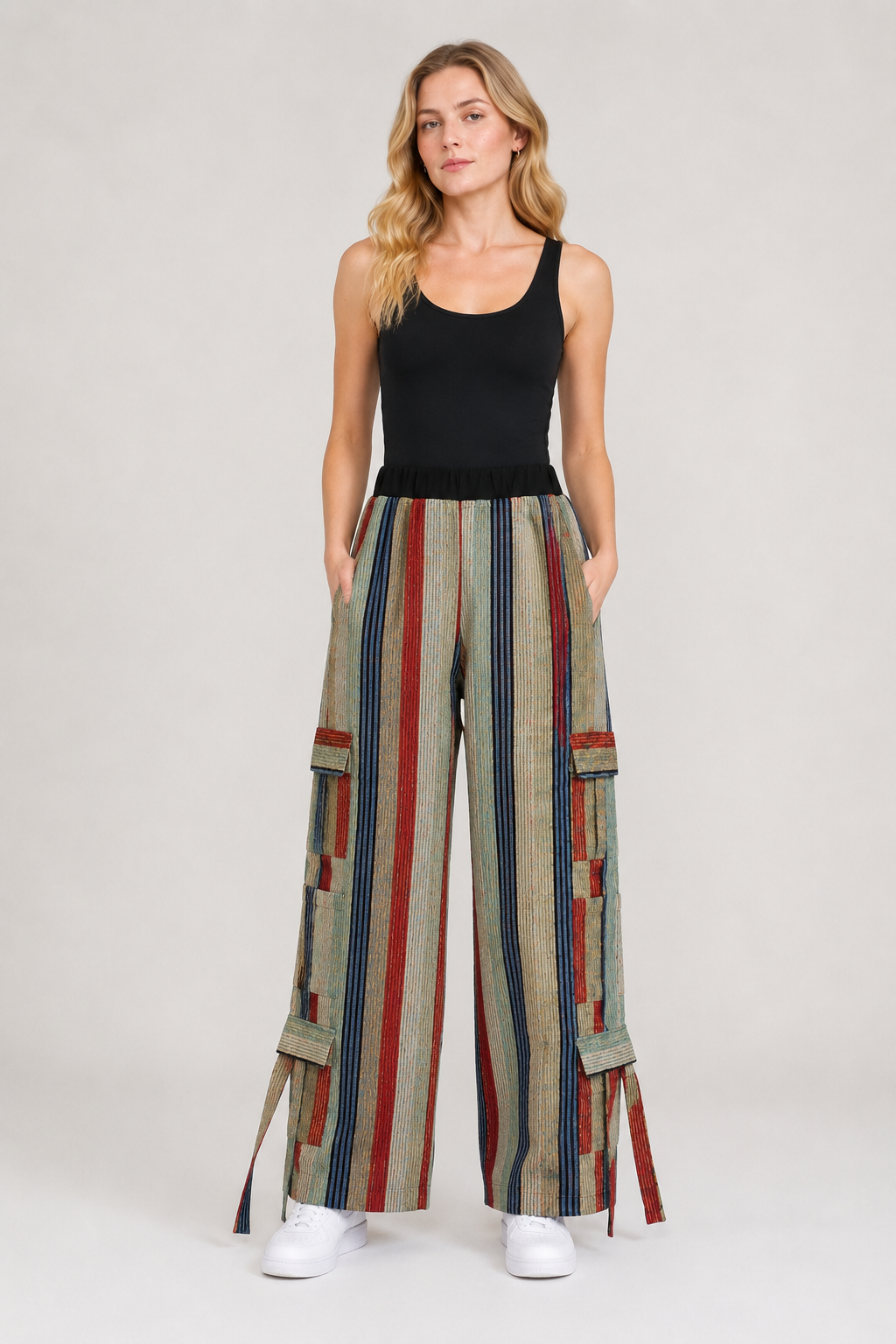 A woman modeling wide-leg cargo pants with vertical stripes in beige, red, blue, and green, paired with a black tank top and white sneakers, against a light gray studio background.