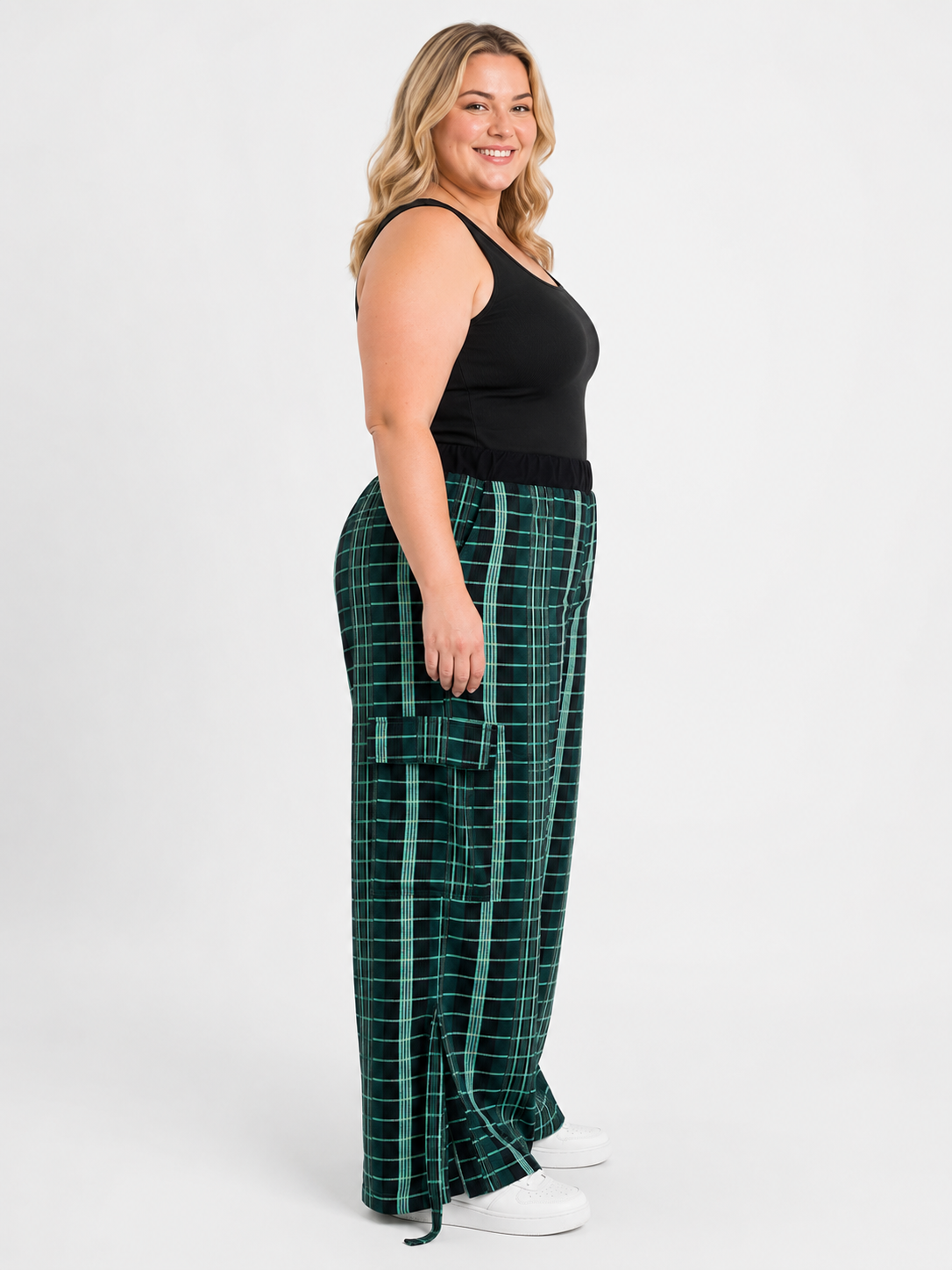 A woman modeling a pair of dark green plaid cargo pants with a black tank top and white sneakers, standing against a light gray studio background.
