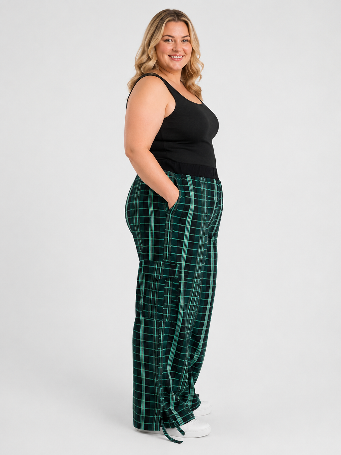 A woman modeling a pair of green plaid lounge pants with a black tank top and white sneakers, standing against a light gray studio background.