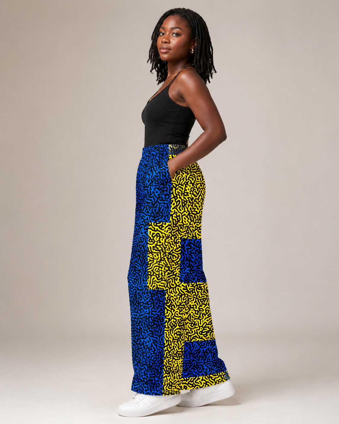A woman modeling a pair of wide-leg pants featuring a bold blue and yellow abstract pattern, paired with a black tank top and white sneakers, set against a plain beige studio background.