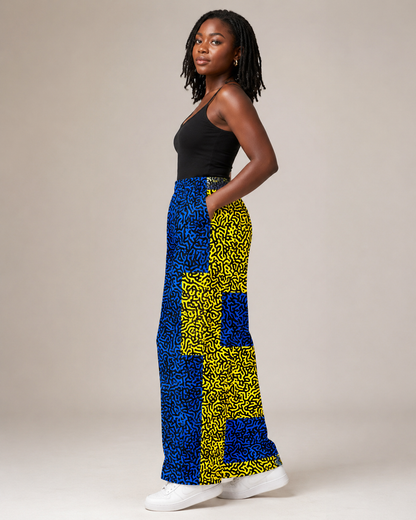 A woman modeling a pair of wide-leg pants featuring a bold blue and yellow abstract pattern, paired with a black tank top and white sneakers, set against a plain beige studio background.
