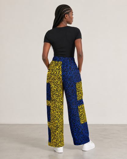 A woman is shown from behind wearing high-waisted wide-leg pants with a bold yellow and blue abstract pattern, paired with a black t-shirt and white sneakers, against a neutral studio background.