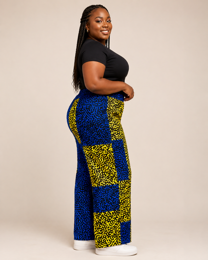A BB woman modeling a pair of wide-leg pants with a bold blue and yellow checkerboard pattern, paired with a black t-shirt and white sneakers, against a solid beige studio background.