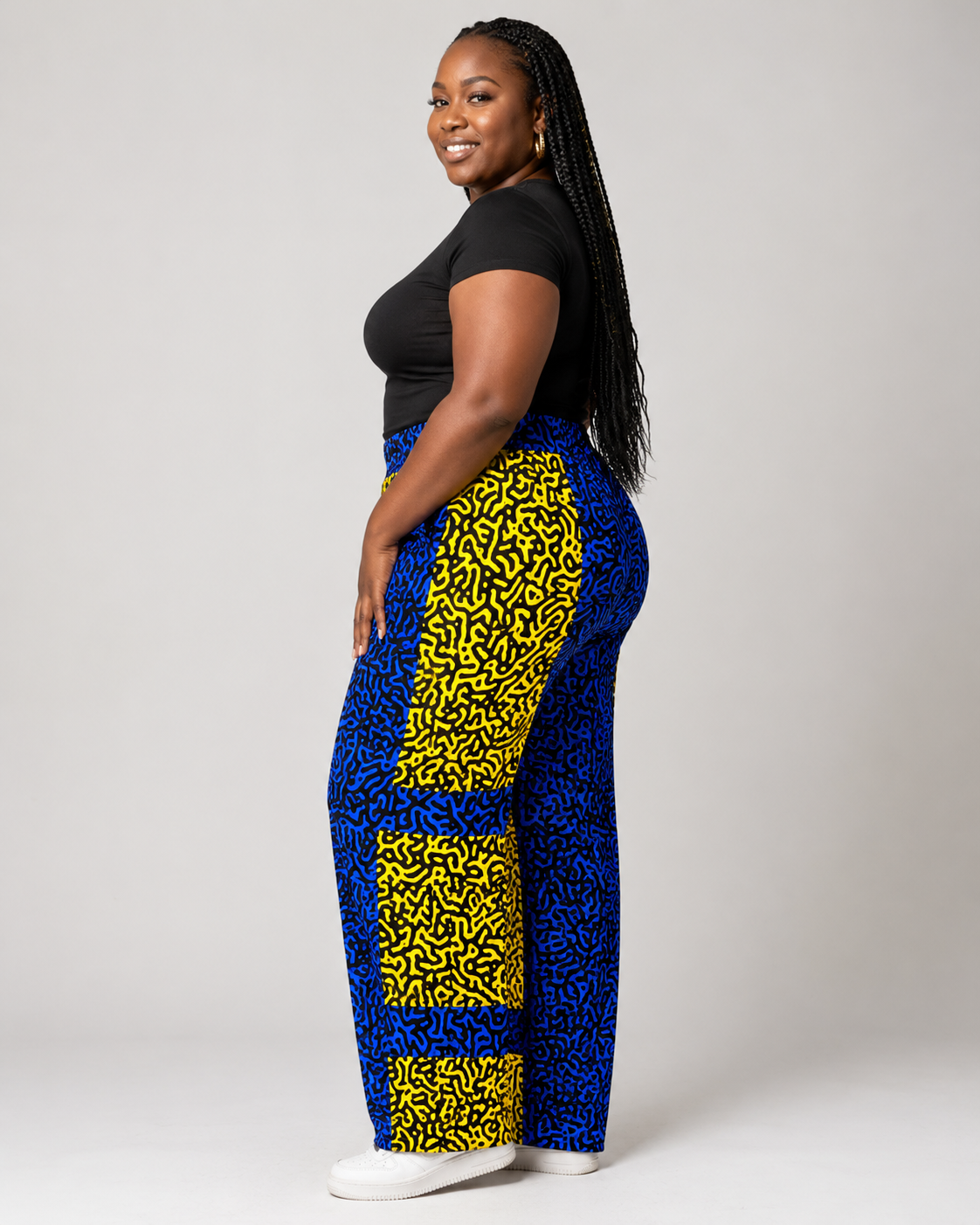 A bold woman modeling high-waisted wide-leg pants with a bold blue and yellow abstract pattern, paired with a black top and white sneakers, against a light gray studio background.
