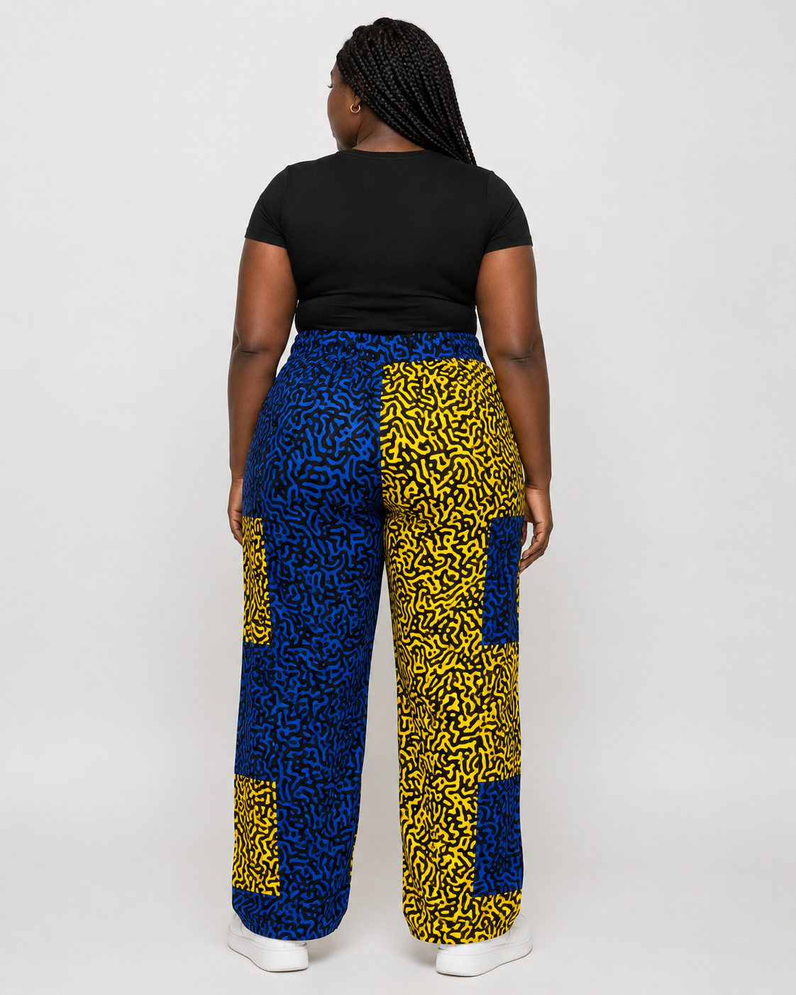 A BB person shown from the back wearing wide-leg pants with a bold blue and yellow abstract pattern, paired with a black t-shirt and white sneakers, against a plain light gray studio background.