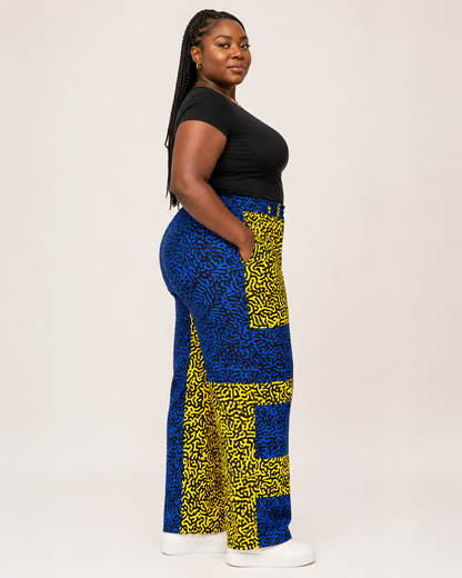 A woman stands in profile wearing a black short-sleeve top and high-waisted, wide-leg pants with a bold blue and yellow abstract pattern. She is posed against a solid, light off-white studio background.