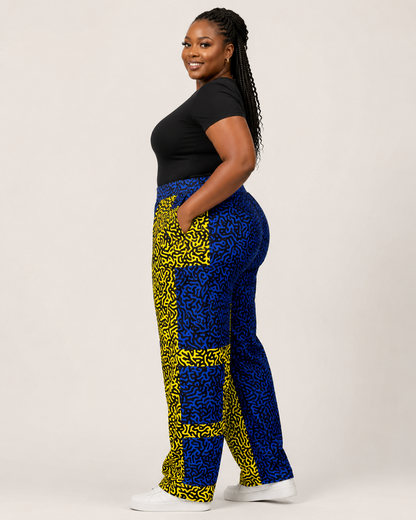 A woman modeling high-waisted, wide-leg pants with a bold blue and yellow abstract pattern, paired with a black top and white sneakers, against a plain light background.