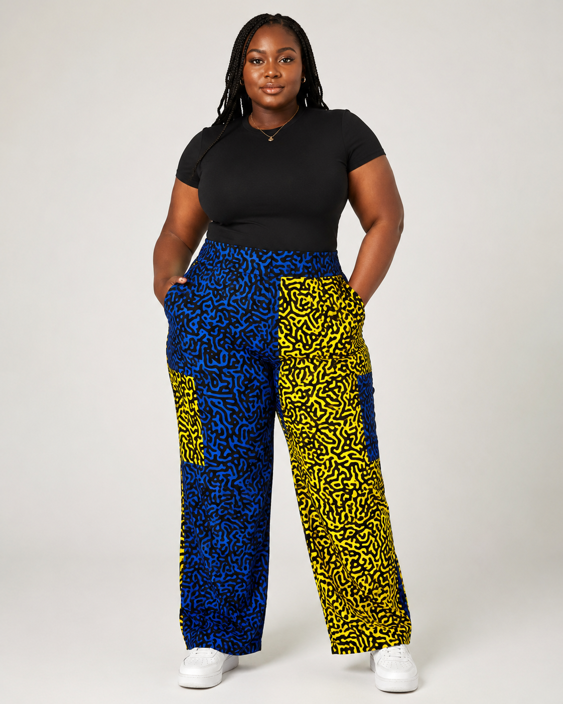 A woman modeling high-waisted, wide-leg pants with a bold blue and yellow abstract pattern, paired with a black t-shirt and white sneakers, against a light gray studio background.