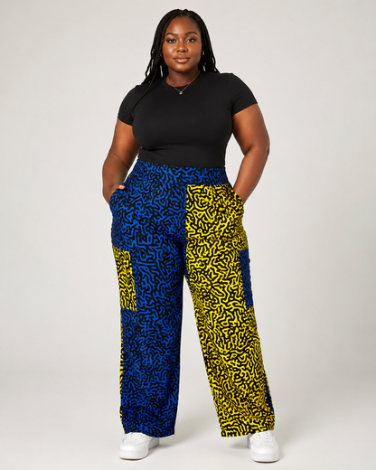 A woman modeling high-waisted, wide-leg pants with a bold blue and yellow abstract pattern, paired with a black t-shirt and white sneakers, against a light gray studio background.