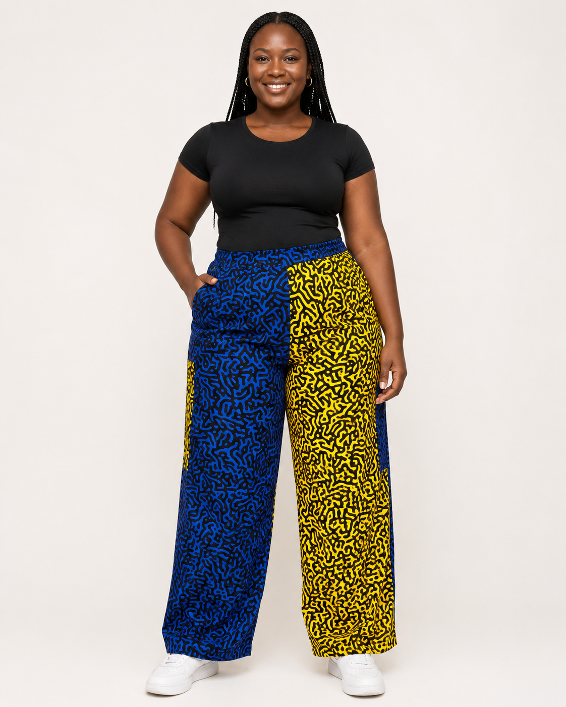 A woman modeling wide-leg pants with a split design: one leg in blue with a black abstract pattern and the other in yellow with a black abstract pattern, paired with a black short-sleeve top and white sneakers against a plain light background.