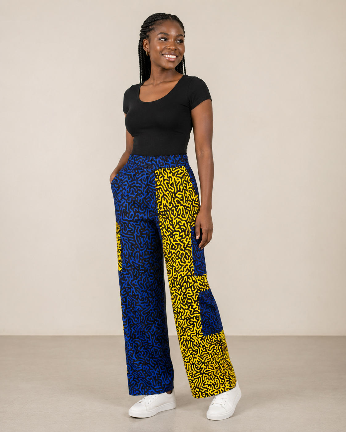 A woman modeling a pair of wide-leg trousers with a bold blue and yellow abstract pattern, paired with a black short-sleeve top and white sneakers, against a plain beige studio background.