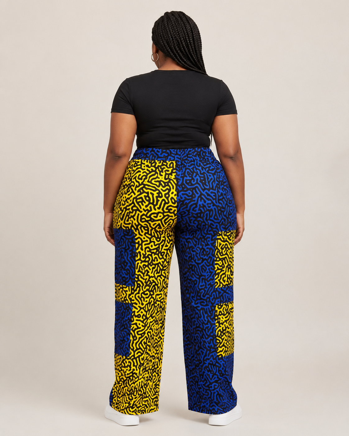 A person shown from the back wearing wide-leg pants with a bold yellow and blue abstract pattern, paired with a black short-sleeve top and white sneakers, against a plain light beige studio background.
