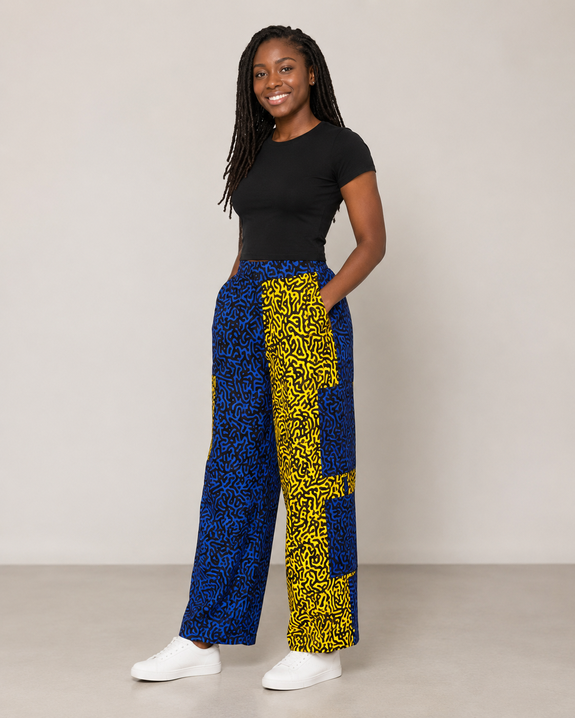 A woman modeling high-waisted wide-leg pants with a bold blue and yellow patterned design, paired with a black t-shirt and white sneakers, standing against a neutral studio backdrop.