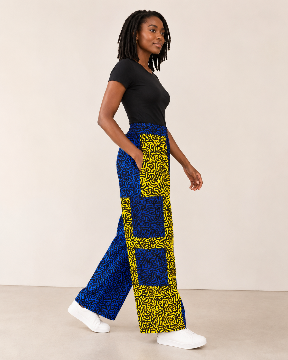 A woman modeling wide-leg pants with a bold blue and yellow geometric pattern, paired with a black t-shirt and white sneakers, set against a plain beige studio background.