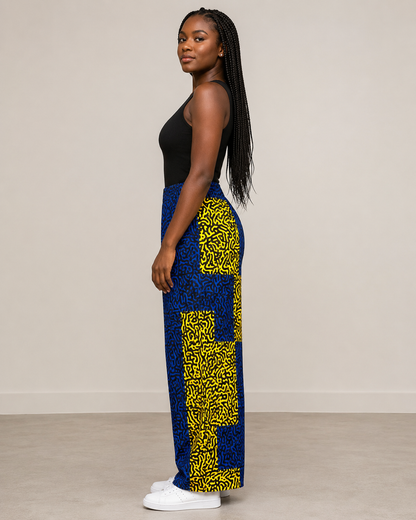 A woman stands in profile wearing a black sleeveless top, blue and yellow patterned wide-leg pants, and white sneakers against a plain beige studio wall and floor.