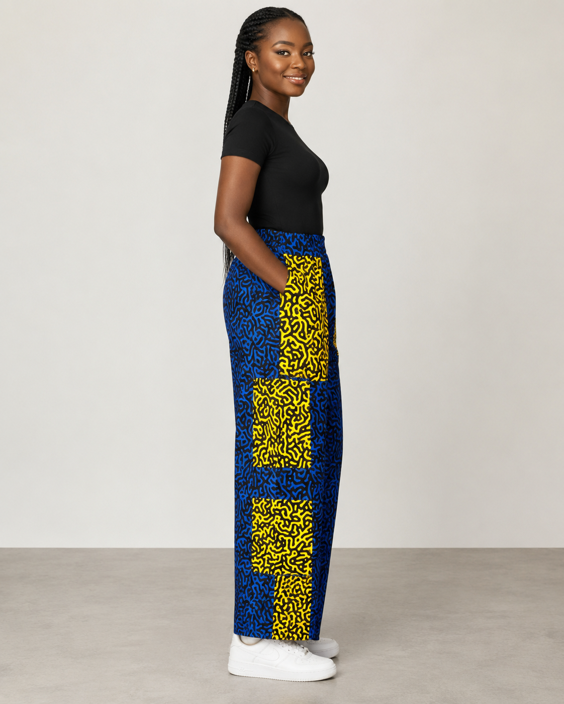 A woman stands in profile wearing a black short-sleeve top and high-waisted wide-leg pants with a bold blue and yellow abstract pattern, paired with white sneakers, against a plain light gray studio background.
