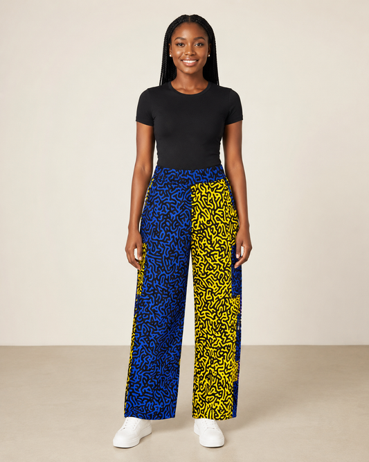 A woman modeling high-waisted wide-leg pants with a split abstract pattern, featuring blue and yellow with black scribble designs, paired with a black t-shirt and white sneakers against a neutral studio backdrop.