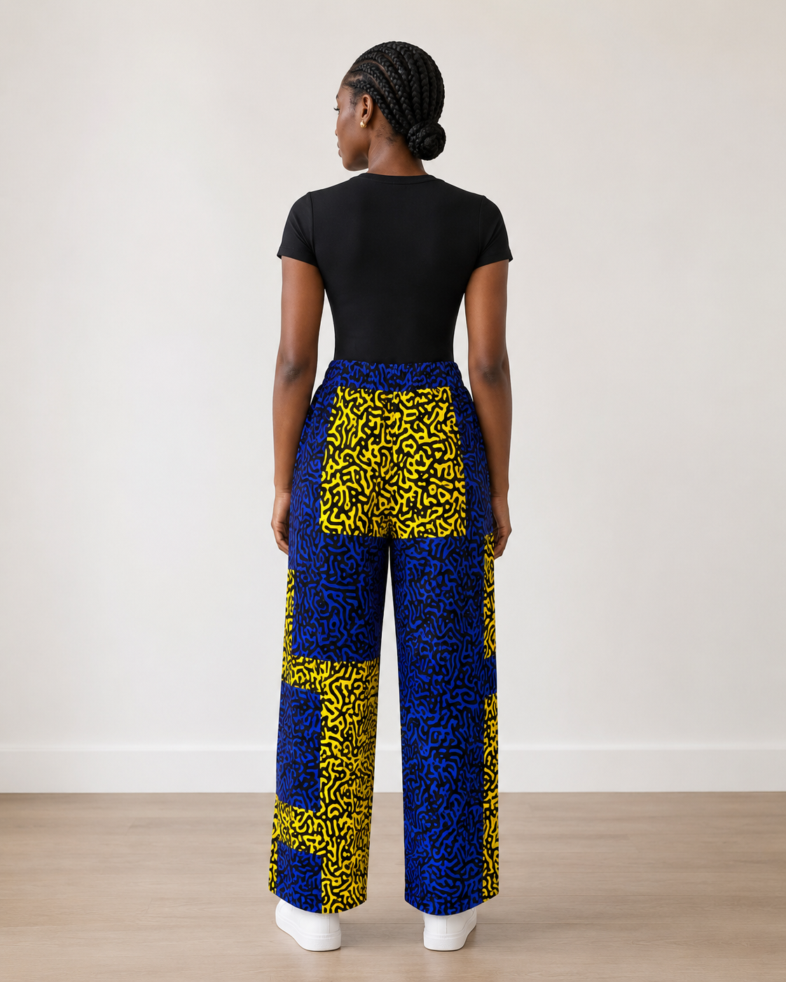 A woman shown from the back wearing a black short-sleeve top and high-waisted wide-leg pants with a bold yellow and blue abstract pattern, standing against a plain off-white wall.