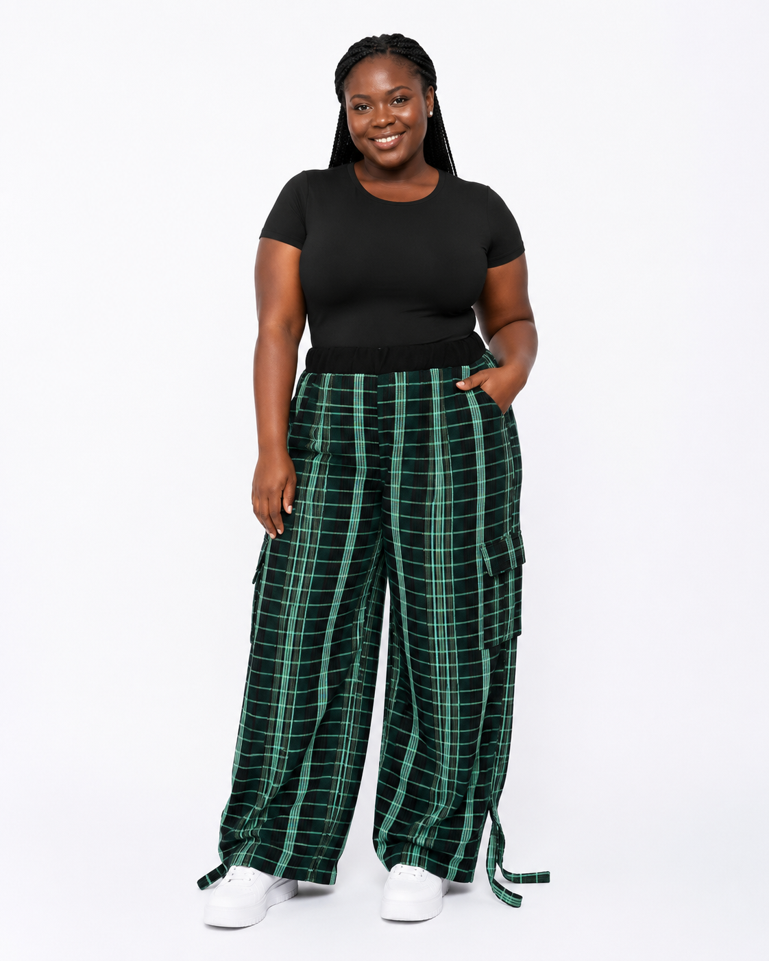 A woman modeling wide-leg green plaid cargo pants with a black t-shirt and white sneakers, standing against a plain white studio background.