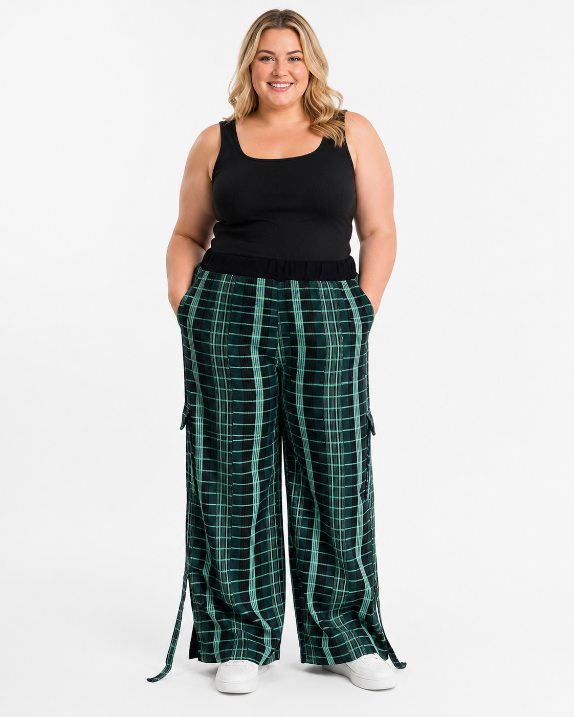 A woman modeling a pair of wide-leg green plaid cargo pants with side pockets and a matching belt, paired with a black tank top and white sneakers, against a plain white studio background.