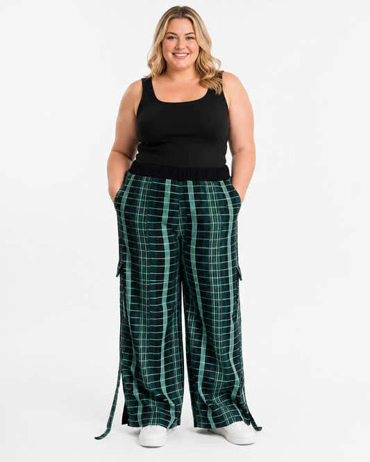 A woman modeling a pair of wide-leg green plaid cargo pants with side pockets and a matching belt, paired with a black tank top and white sneakers, against a plain white studio background.