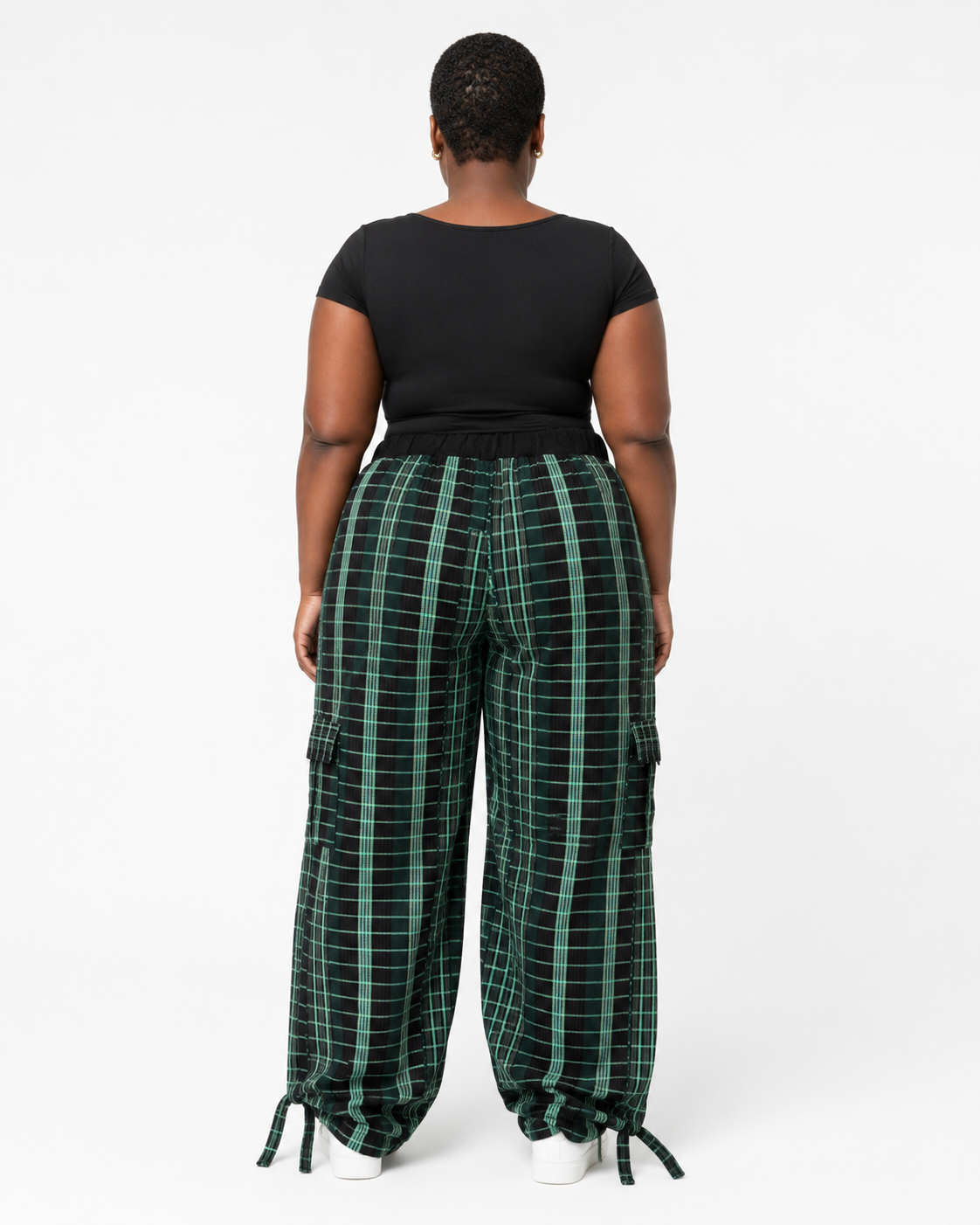 A model viewed from the back wearing wide-leg green and black plaid cargo pants with a black short-sleeve top, set against a plain white studio background.