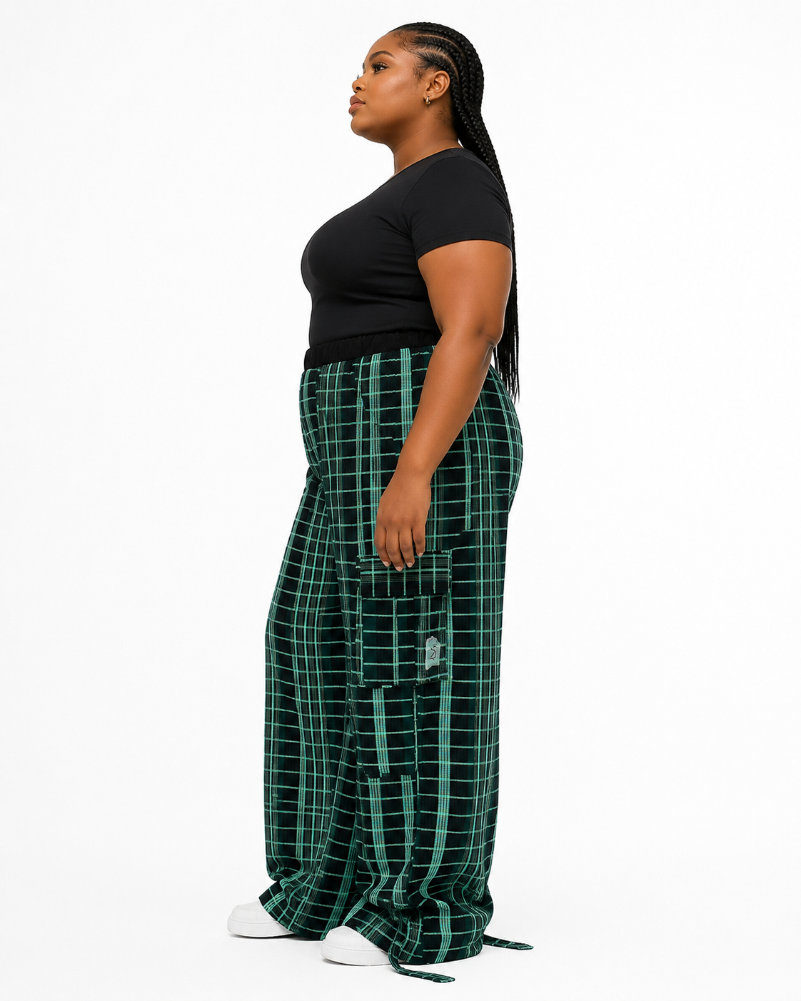 A woman in a black short-sleeve top and green plaid cargo pants stands in profile against a plain white studio background.