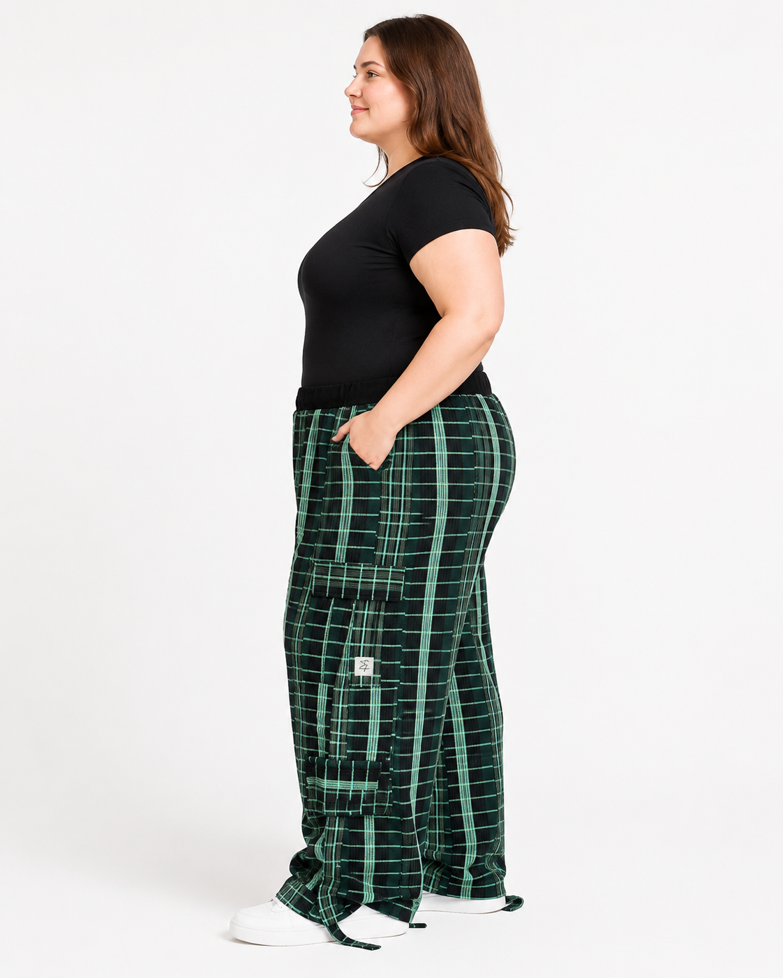 A woman stands in profile wearing dark green and black plaid cargo pants, a black short-sleeve top, and white sneakers against a plain white studio background.